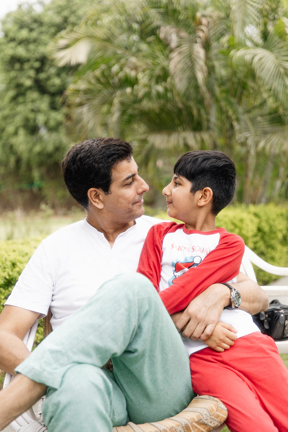 Father and son sharing a loving moment outdoors, sitting together on a bench and smiling warmly at each other in a lush garden setting