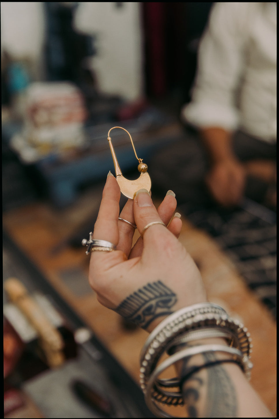 Main tatouée ornée de bagues et bracelets tenant une boucle d’oreille artisanale dorée en forme de croissant avec perle décorative.