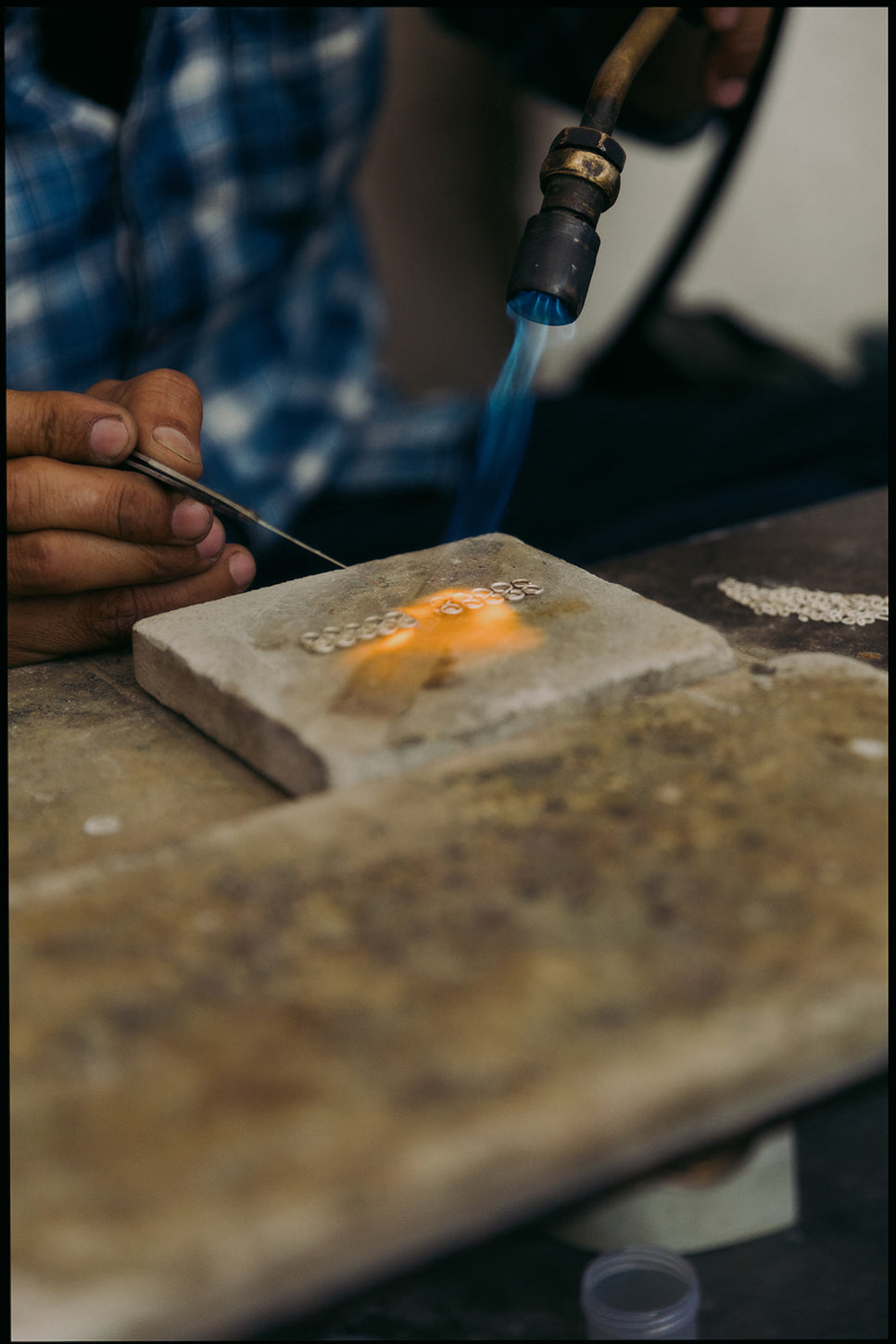Artisan en chemise bleue soudant des anneaux métalliques à l’aide d’un chalumeau, avec flamme bleue et orange sur pierre réfractaire.
