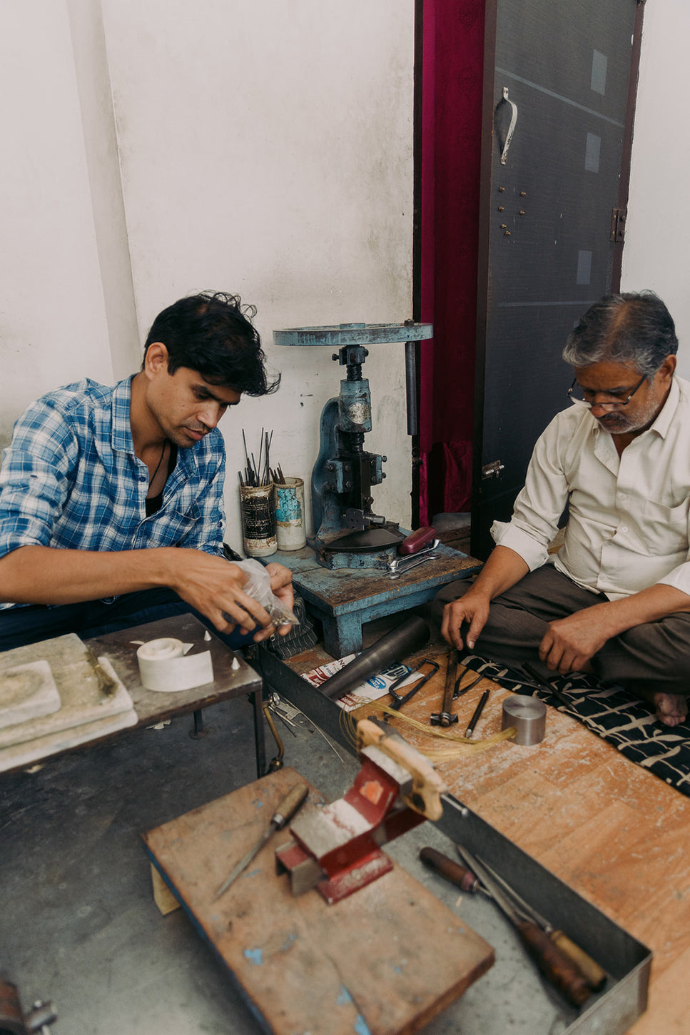 Two artisans working with hand tools in a small artisan workshop, surrounded by metalworking equipment and traditional crafting materials.