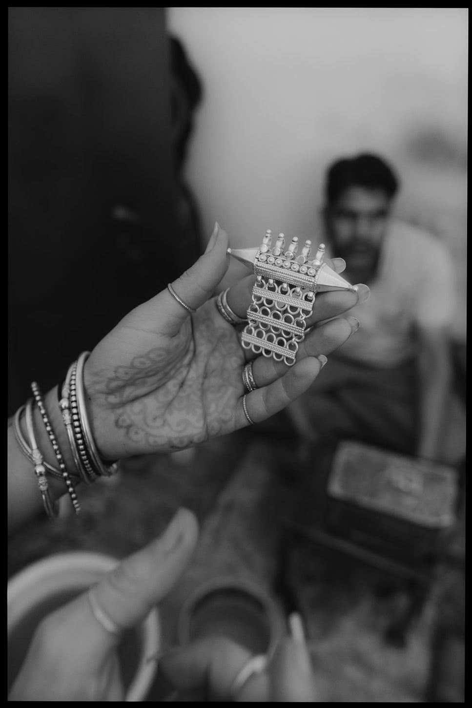 Close-up of a woman’s henna-decorated hand holding a detailed piece of handmade jewelry, with a blurred artisan in the background.