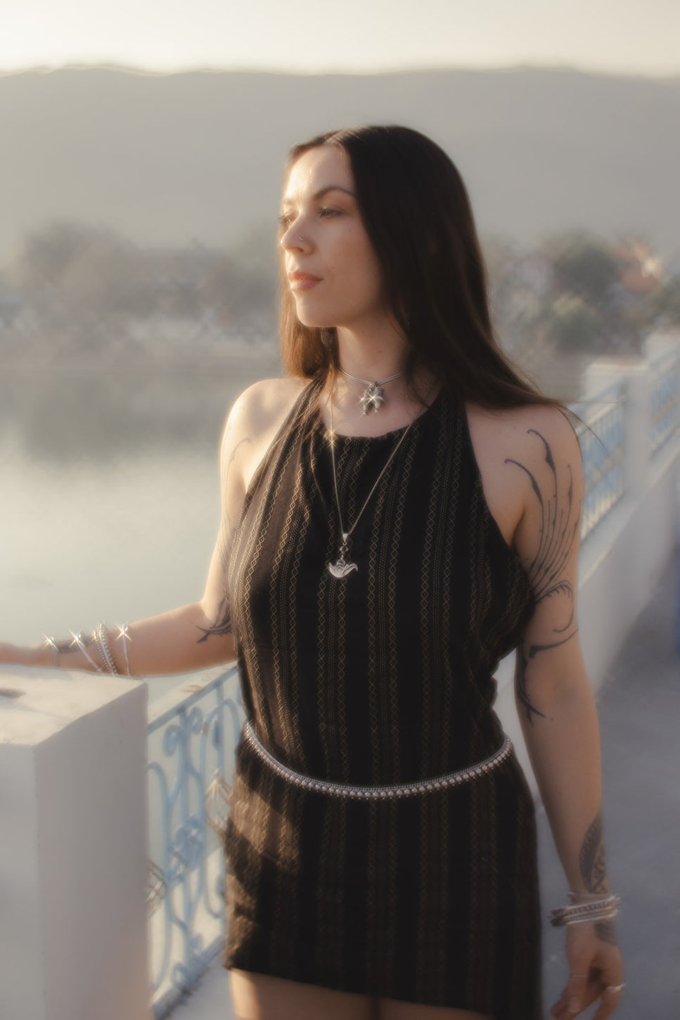 Woman standing outdoors in soft sunlight, wearing layered silver jewelry and a black sleeveless dress, with tattoos on her arm, looking serene by the water.