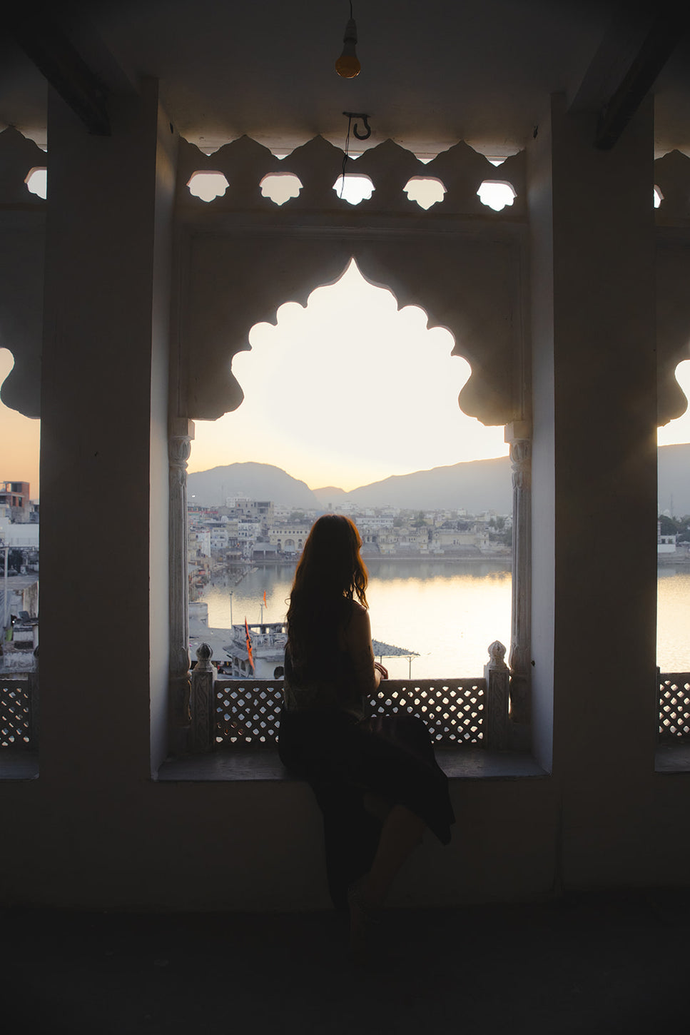 Silhouette of a woman gazing at a peaceful sunset view over a lake and cityscape, framed by traditional Indian architecture with scalloped arches.