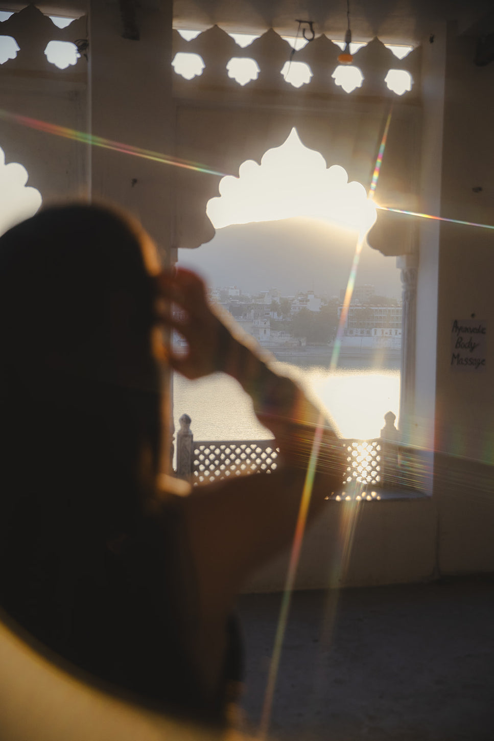 Silhouette floue d’une femme devant une arche traditionnelle au coucher du soleil, avec vue sur un lac et des collines illuminées.
