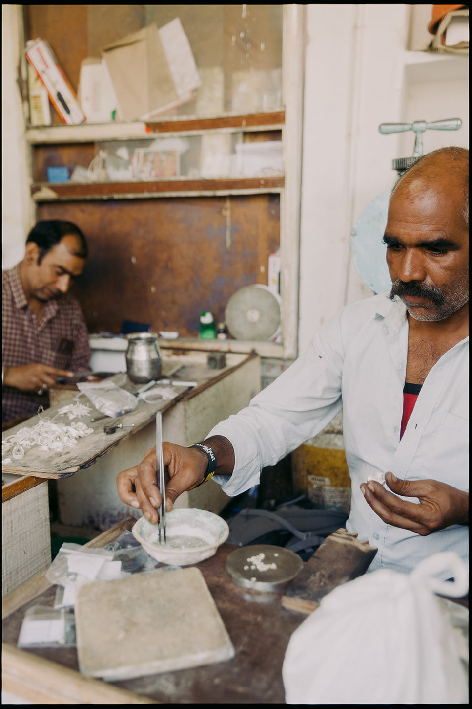 Two artisans work at a jewelry workshop table, one using tweezers with silver material while the other focuses on detailed crafting in the background.