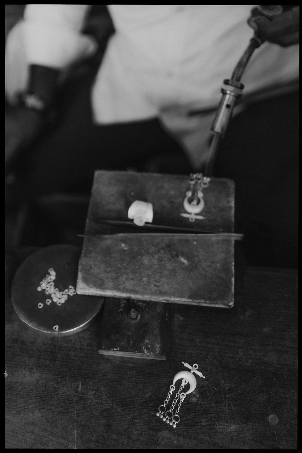 Close-up black and white photo of a jewelry maker soldering a metal piece on a workbench, with small silver elements scattered nearby.