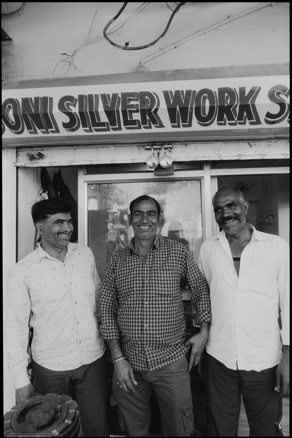 Three men standing and smiling in front of a shop with a sign reading “SILVER WORK,” captured in black and white, conveying a warm, candid moment.