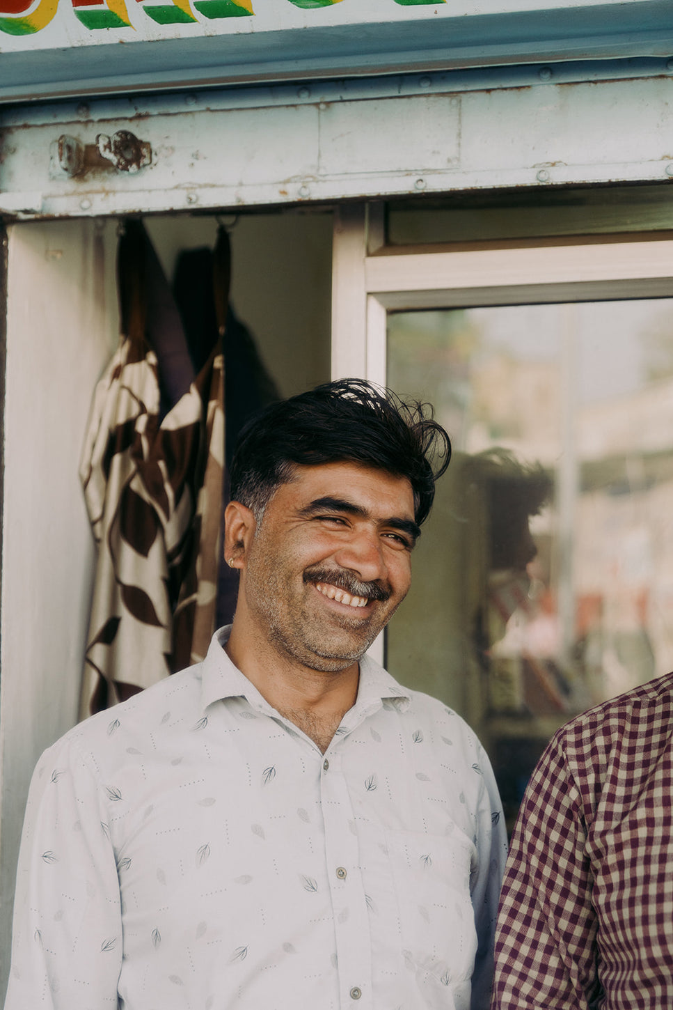 Smiling man in a patterned shirt standing outside a small shop, captured in natural light with a warm, friendly expression symbolizing a local shopkeeper.