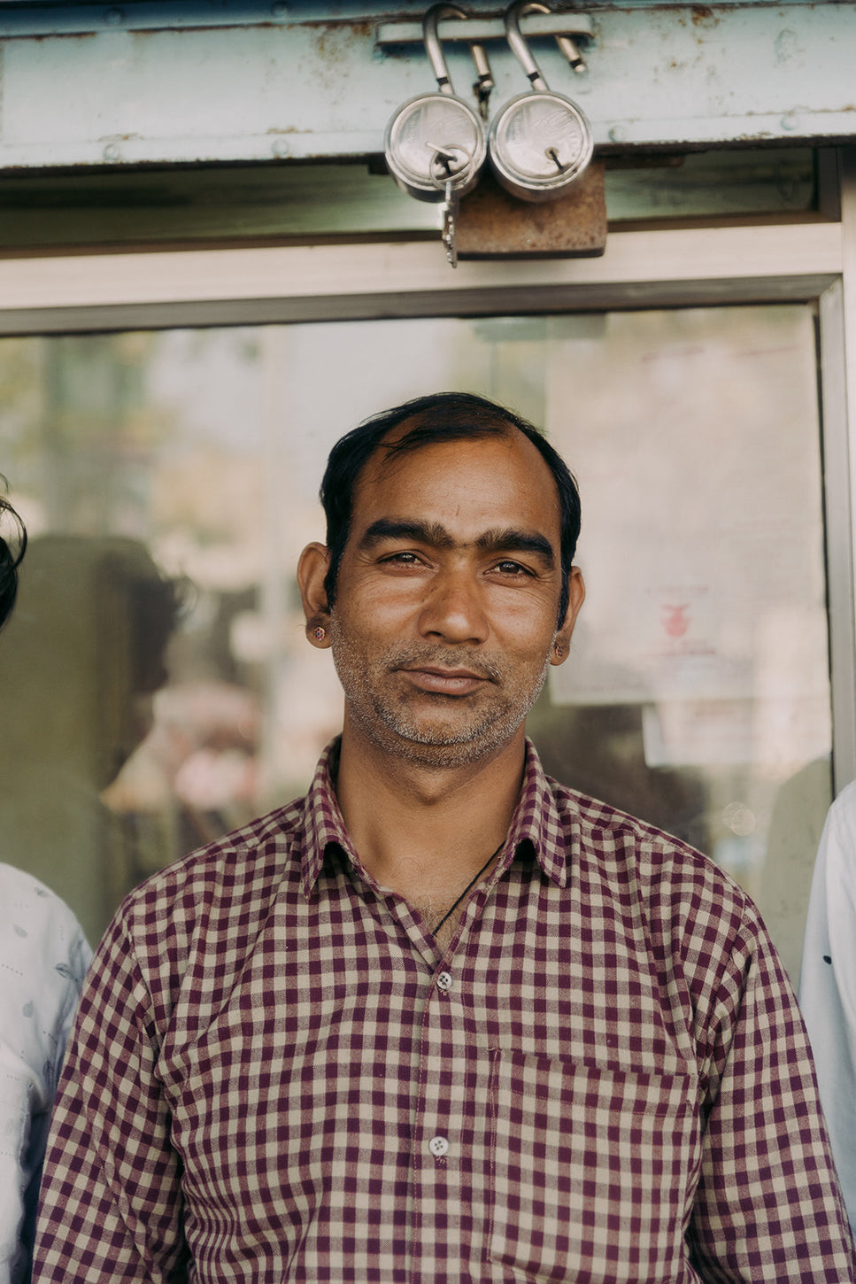 Middle-aged man in a checkered shirt standing confidently in front of a shop window, representing a local entrepreneur or small business owner.