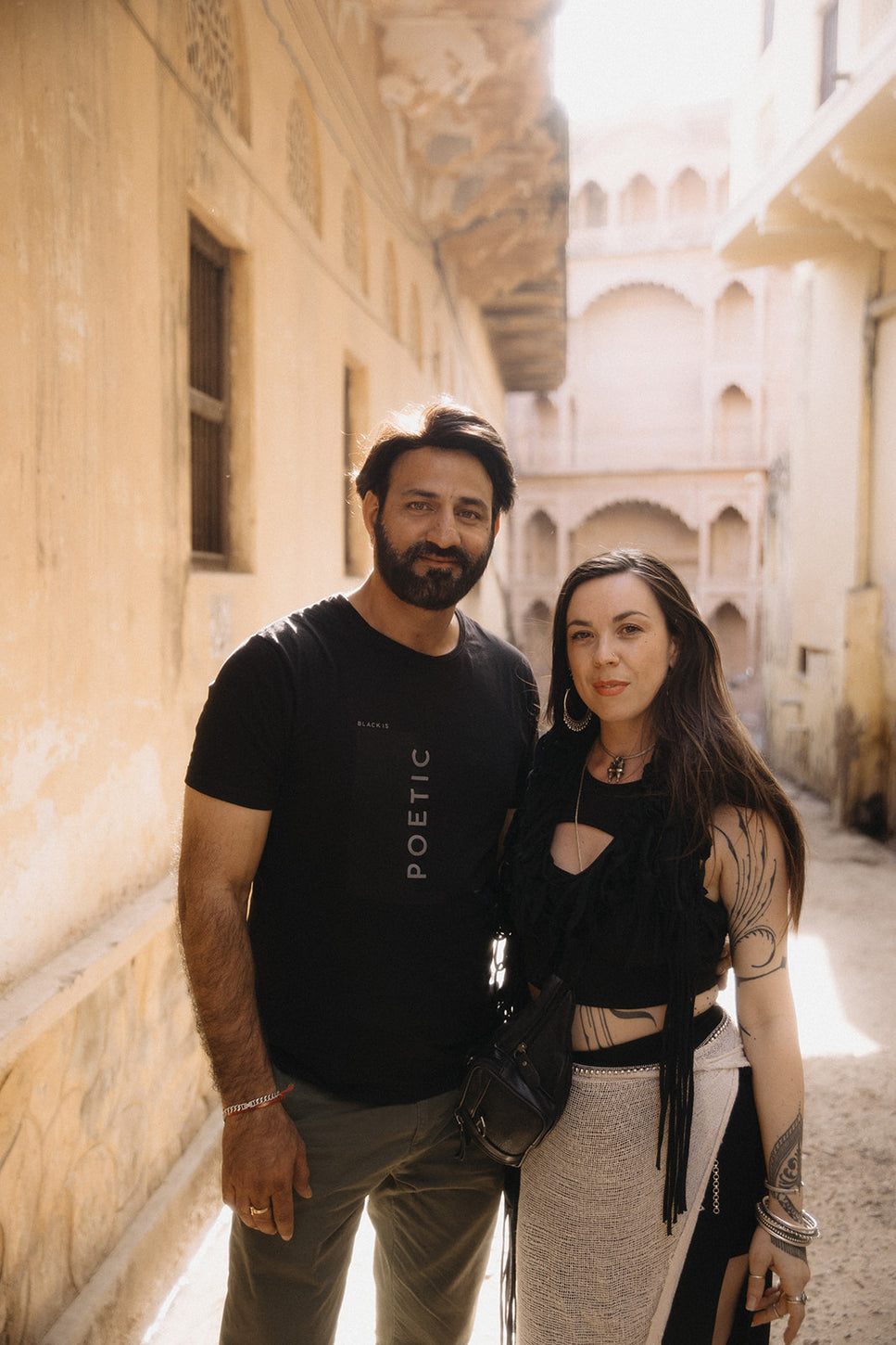 A man and woman standing side by side in a sunlit architectural alley with traditional arches, both wearing black outfits and jewelry.
