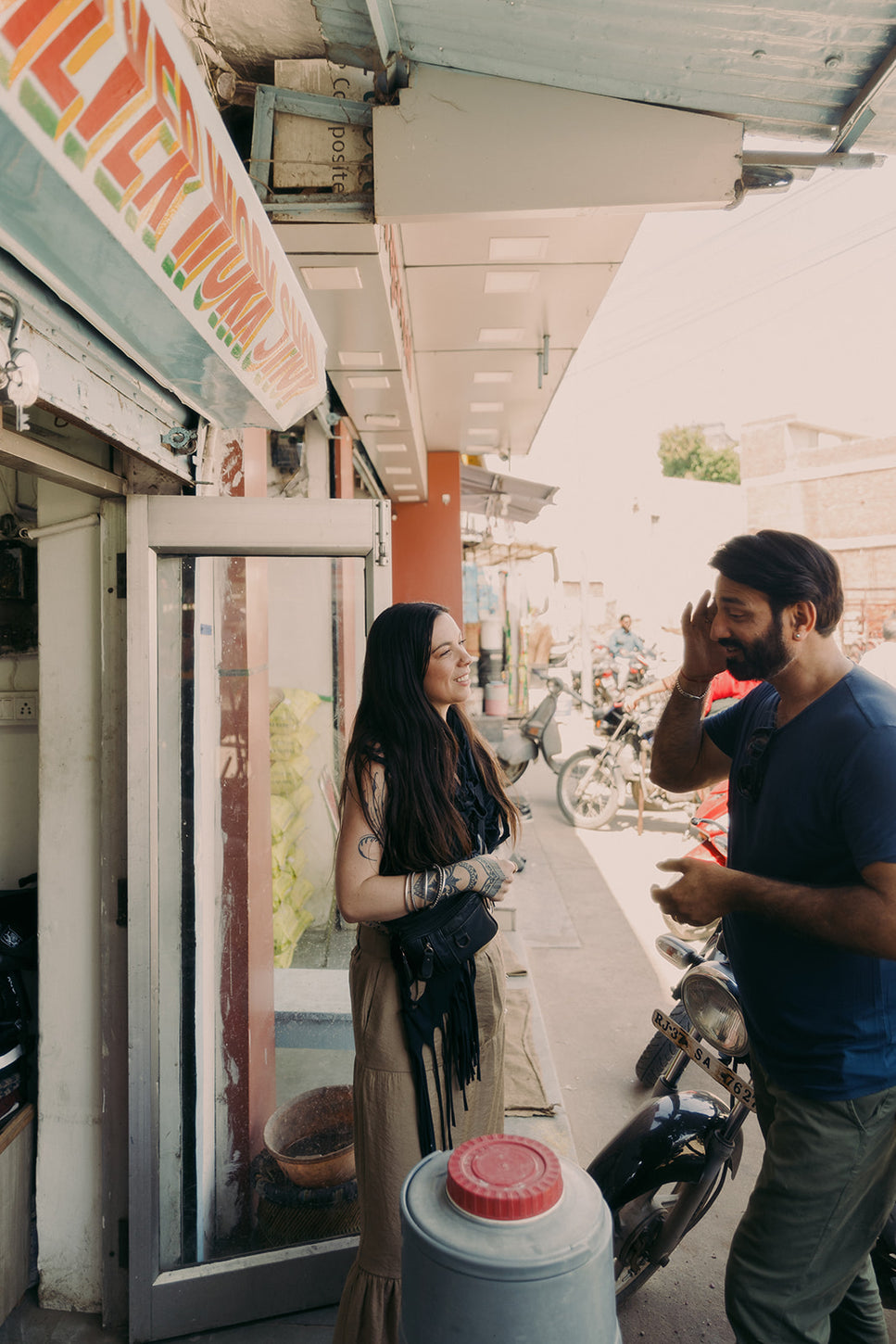 Two people sharing a lighthearted street market conversation outside a local shop, surrounded by scooters and vibrant neighborhood activity.