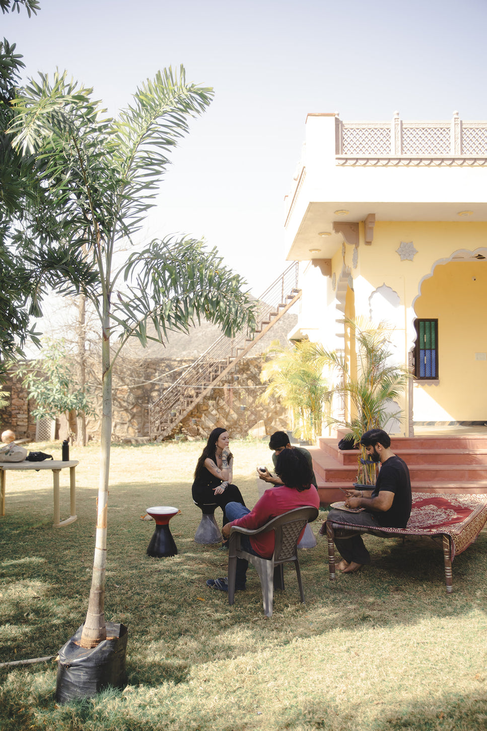 Small group engaged in a relaxed outdoor discussion near a yellow villa, seated on chairs and a charpai under sunny skies and lush greenery.
