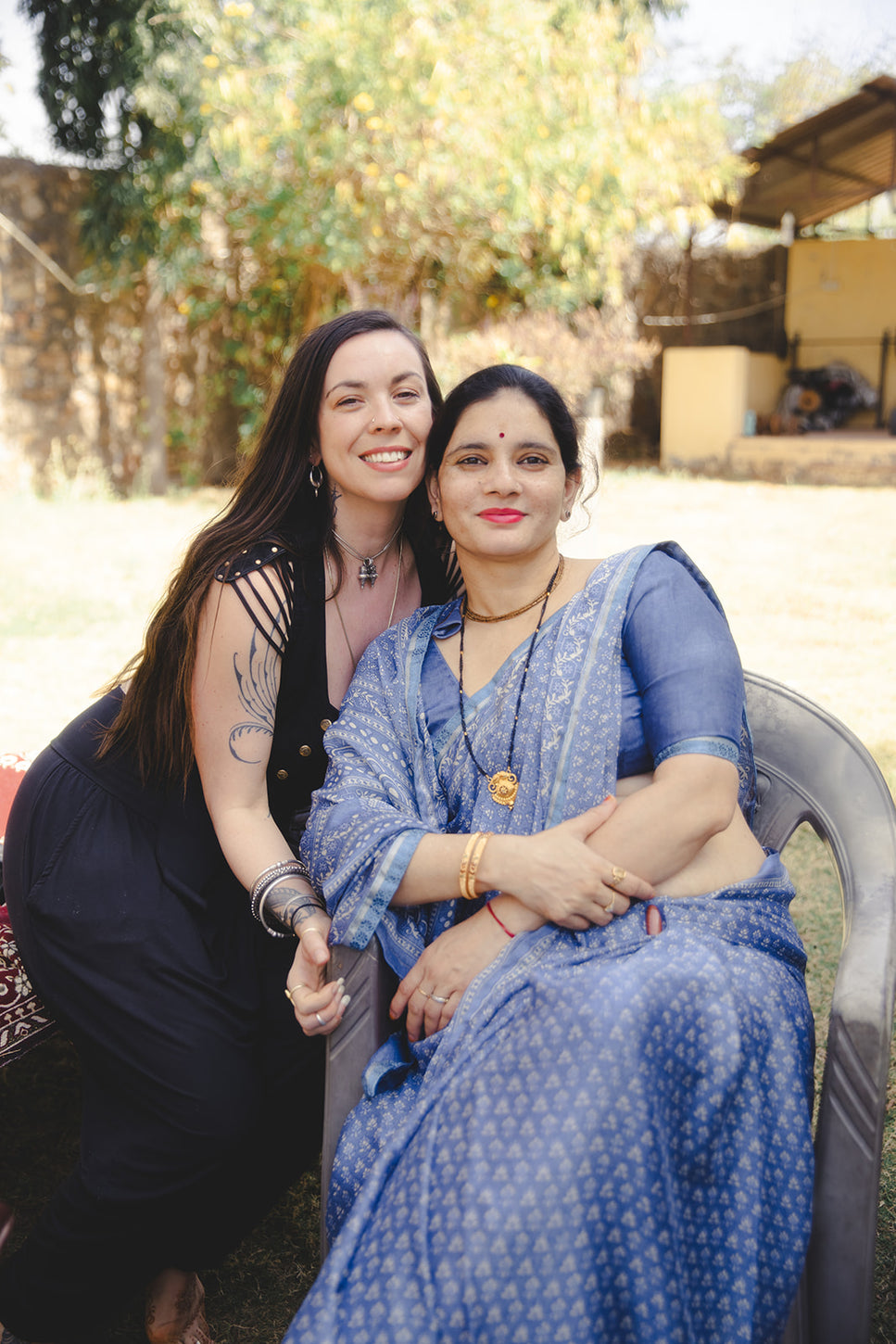 Two women smiling outdoors, one in a black dress with jewelry, the other in a blue saree, sitting close together on a chair.