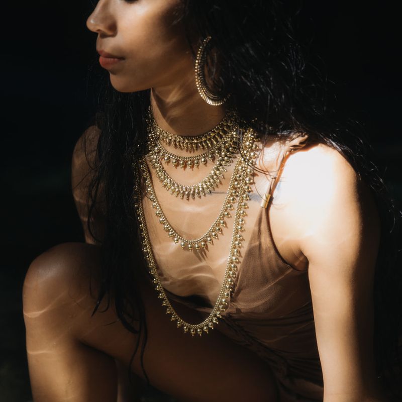 Model wearing stacked layered gold necklaces featuring a beaded fringe choker and crescent hoop earrings against a dark studio backdrop.