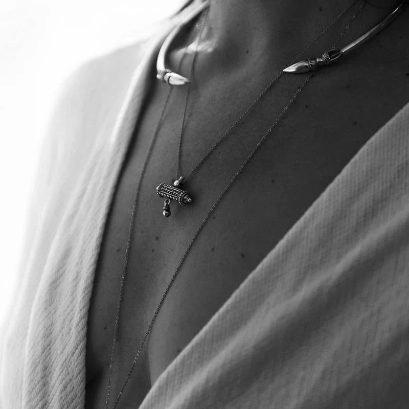 Layered silver pendant necklaces worn on a model's décolletage, featuring a barrel amulet charm on a fine chain with a beaded link collar necklace layered above on 925 sterling silver.
