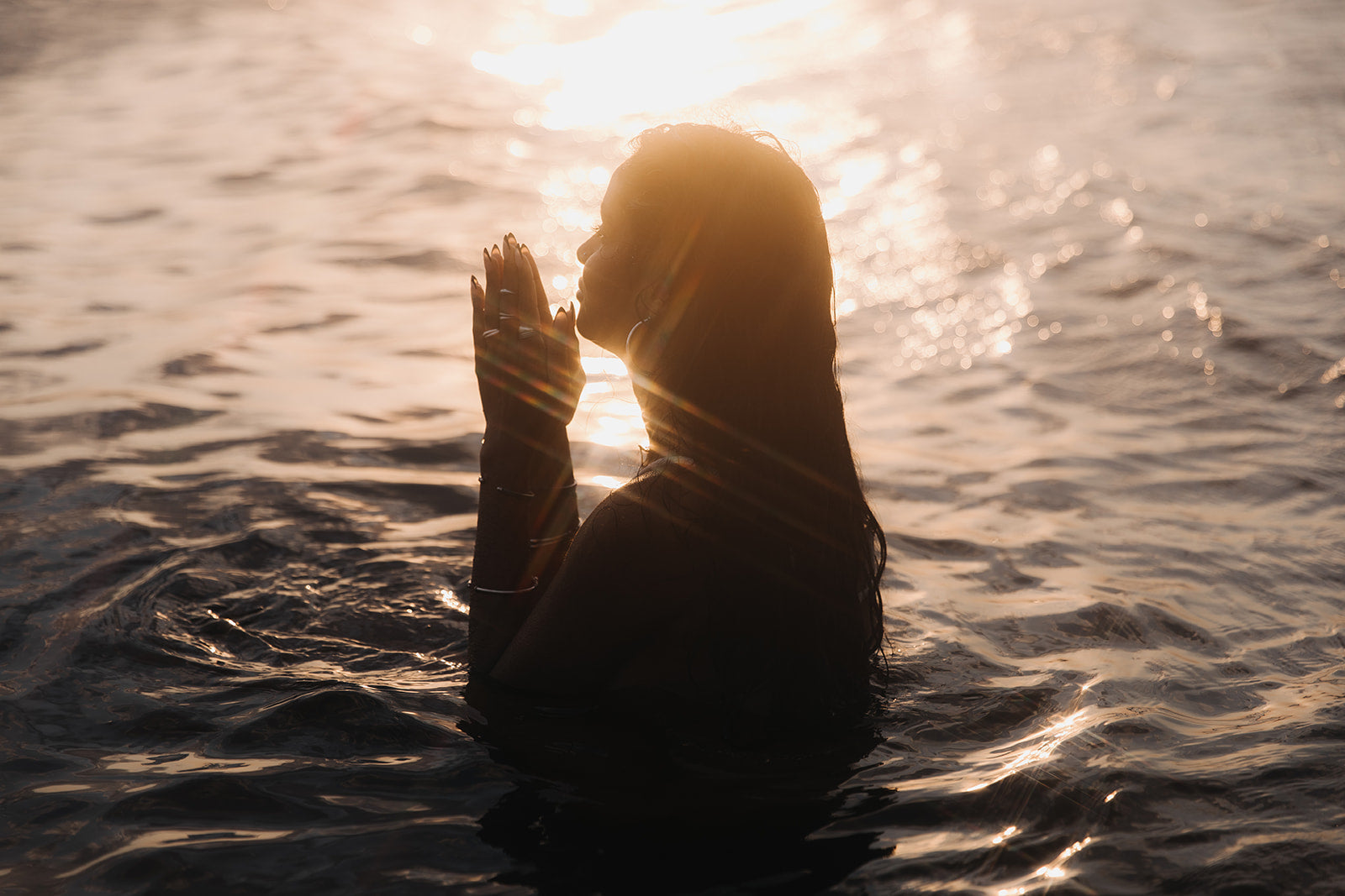 Woman standing in water at sunset, hands raised near her face, wearing Noir KĀLA silver jewelry that catches the warm, shimmering light.