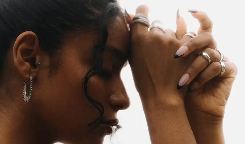 Close-up side profile of a woman with hands pressed together in a prayer pose, wearing Noir KĀLA silver rings and hoop earrings.